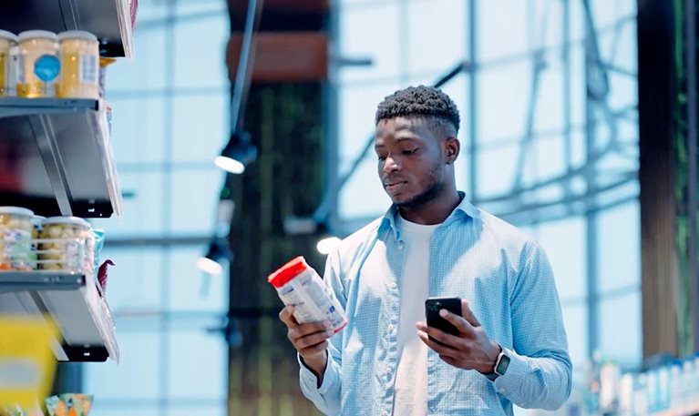 Person in a grocery store comparing a product label while holding a smartphone next to a shopping cart.