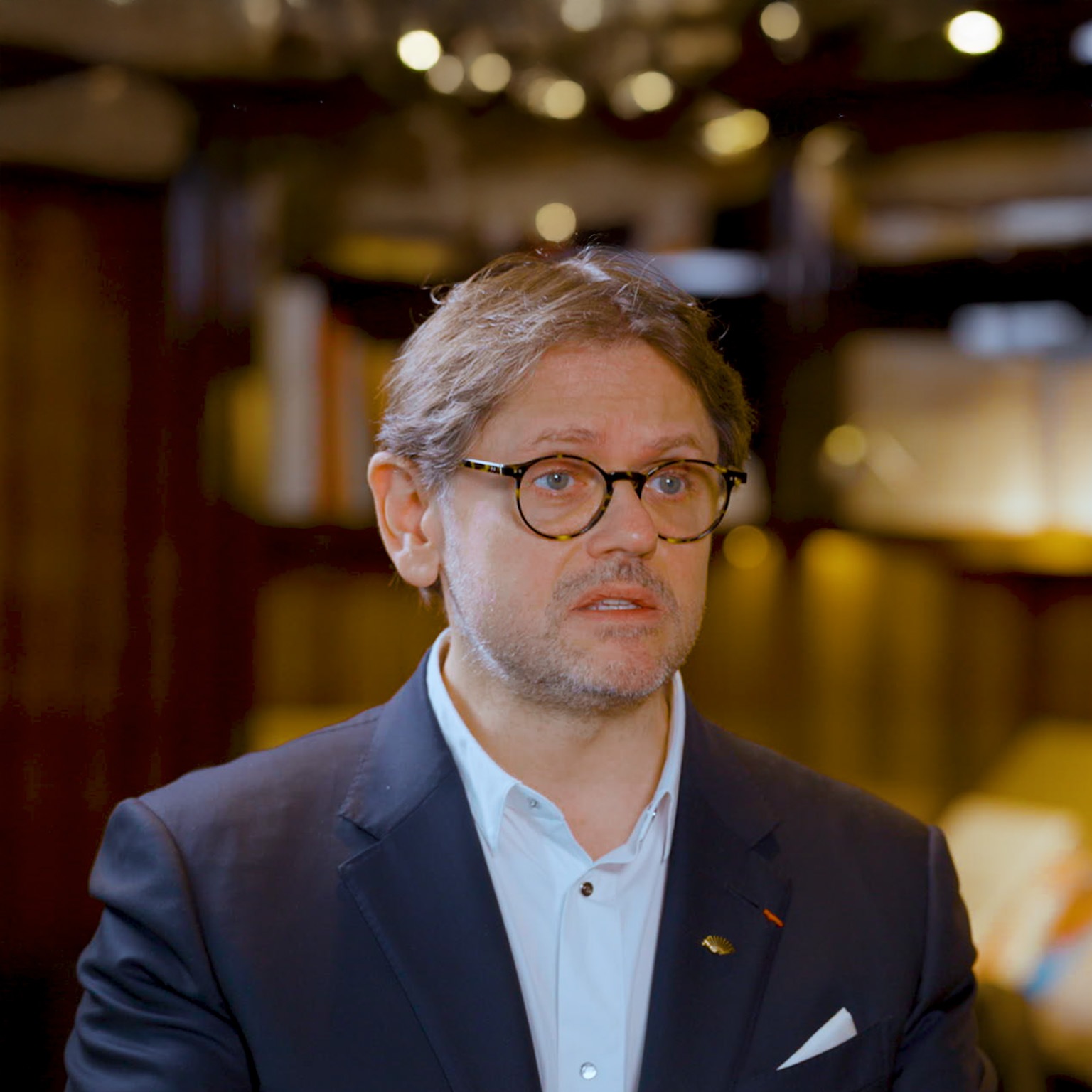 Laurent Kleitman wearing round glasses, a navy blazer, and a light shirt, speaking to Alex Sawaya during an interview. 