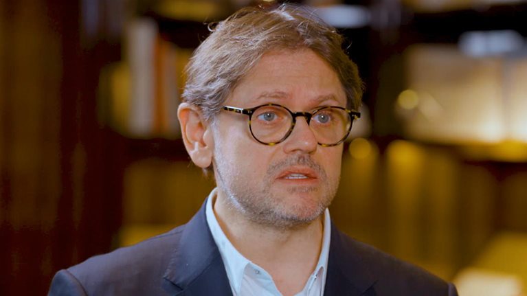 Laurent Kleitman wearing round glasses, a navy blazer, and a light shirt, speaking to Alex Sawaya during an interview.