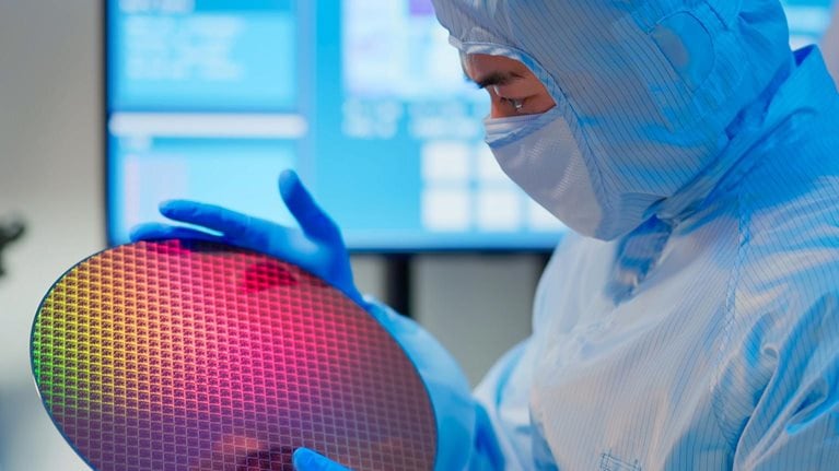 Male technician in sterile coverall holds wafer that reflects many different colors with gloves and check it at semiconductor manufacturing plant.