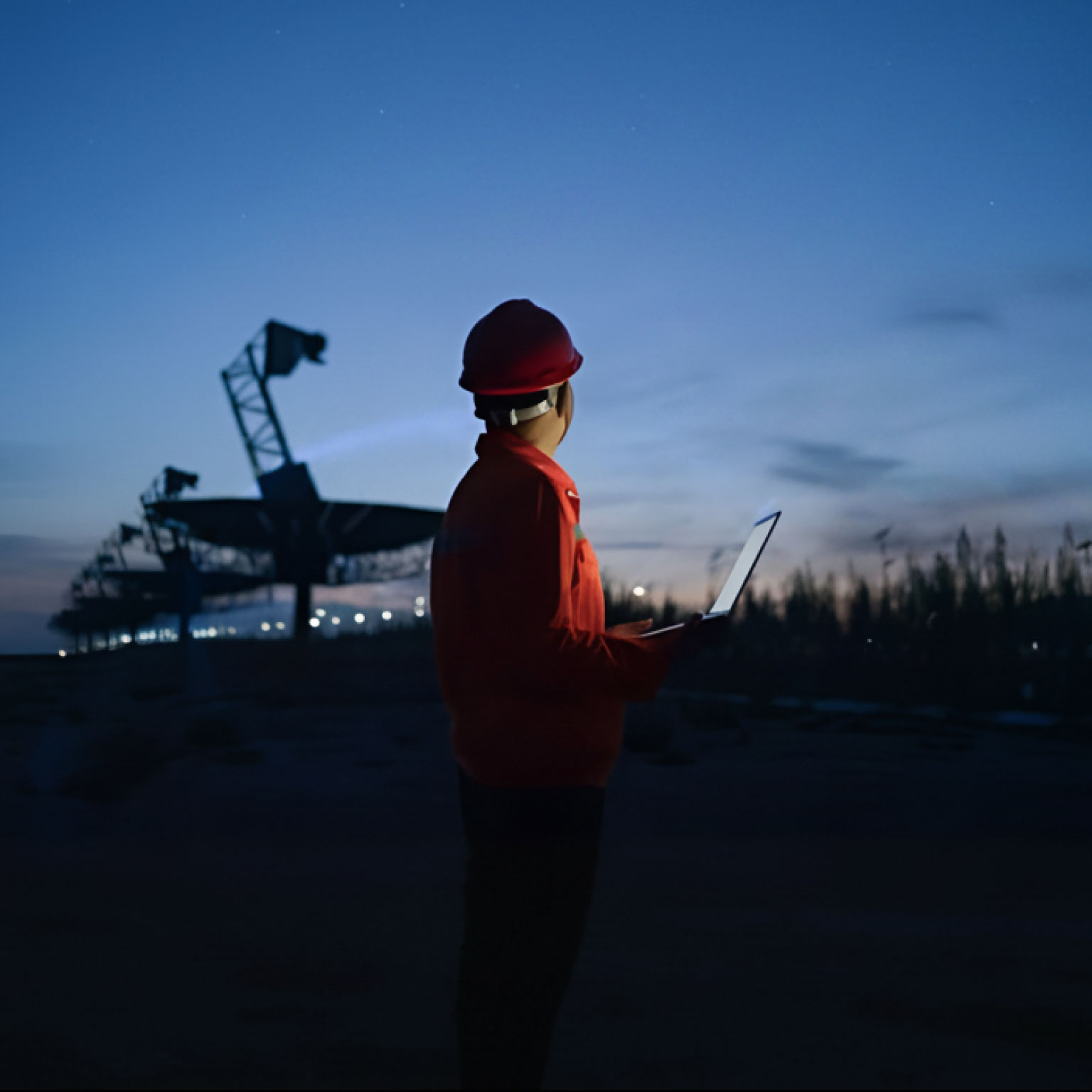 Photograph of a person wearing a red safety helmet and jacket holding a laptop, standing outdoors at dusk near large telecommunications equipment. The scene highlights a work environment with artificial lighting and a darkening sky, suggesting monitoring or inspection activities during low light conditions.