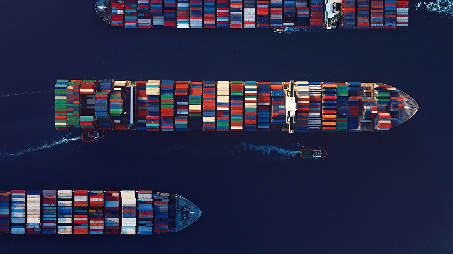 Aerial view of large container ships traveling across deep blue water, stacked with colorful shipping containers arranged in neat rows. Two small boats, highlighted in red, move alongside one of the ships, leaving white wake trails behind them.