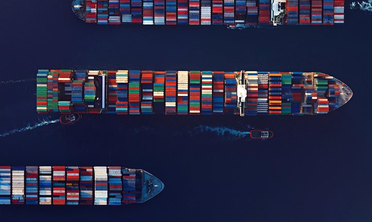 Aerial view of large container ships traveling across deep blue water, stacked with colorful shipping containers arranged in neat rows. Two small boats, highlighted in red, move alongside one of the ships, leaving white wake trails behind them.