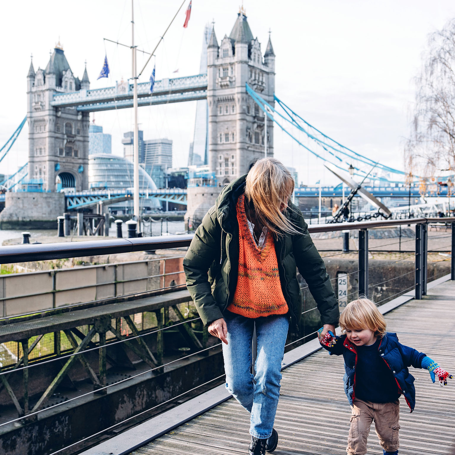 Grandmother holding her grandson's hand while walking on a wooden walkway near the Tower Bridge in London, UK