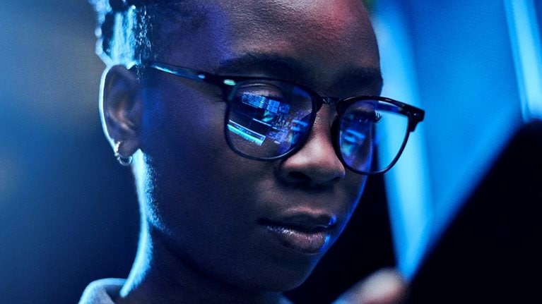 Engineer using a digital tablet while working in a server room with code reflected in her glasses.