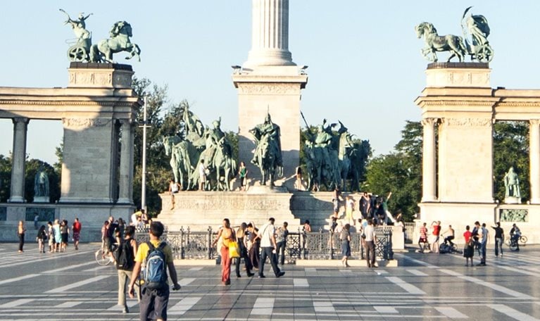 Visitors strolling and capturing images at Heroes Square in Budapest, with the Millennium column prominently displayed at the center of the view.