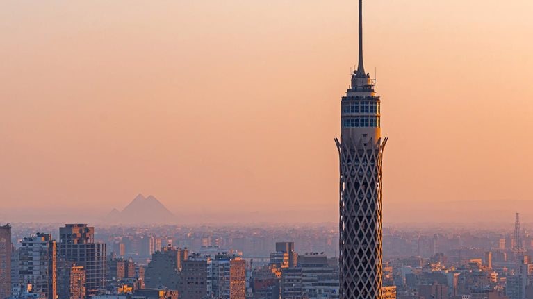 In the golden glow of sunset, Cairo Tower rises above Gezira Island while, in the hazy distance, a Great Pyramid of Giza faintly appears—bridging ancient history and modern Cairo in one quintessential view.