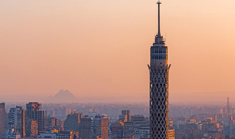In the golden glow of sunset, Cairo Tower rises above Gezira Island while, in the hazy distance, a Great Pyramid of Giza faintly appears—bridging ancient history and modern Cairo in one quintessential view.