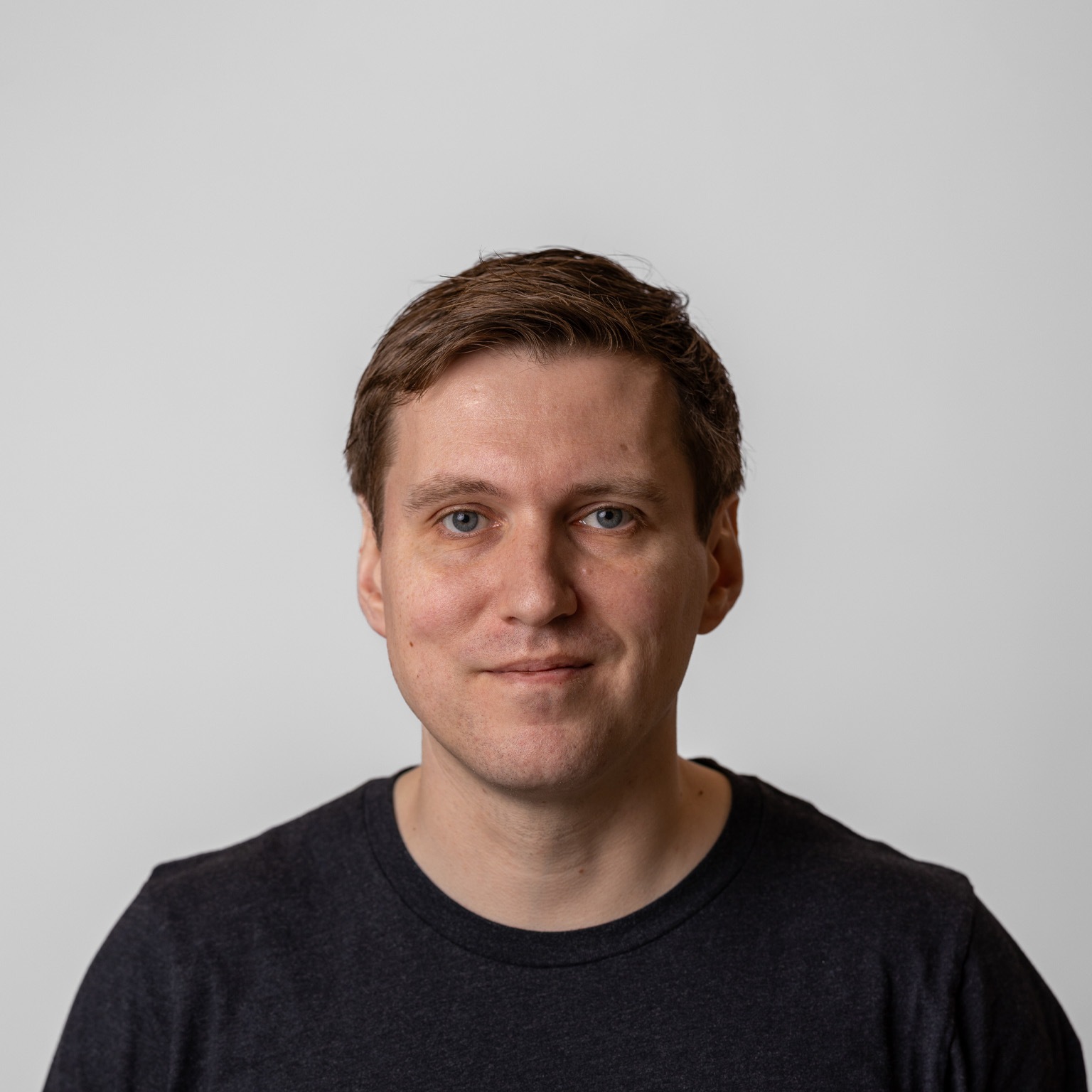 Philip Johnston with short brown hair and blue eyes looks directly at the camera with a slight, relaxed smile. He wears a dark shirt and is set against a plain, light-colored background.