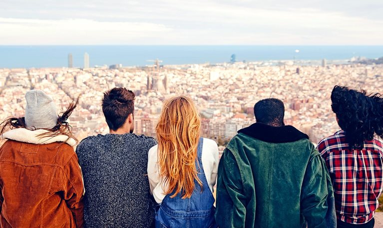 Image from behind of a group of friends sitting on a terrace looking out over a city with the ocean in the far distance.