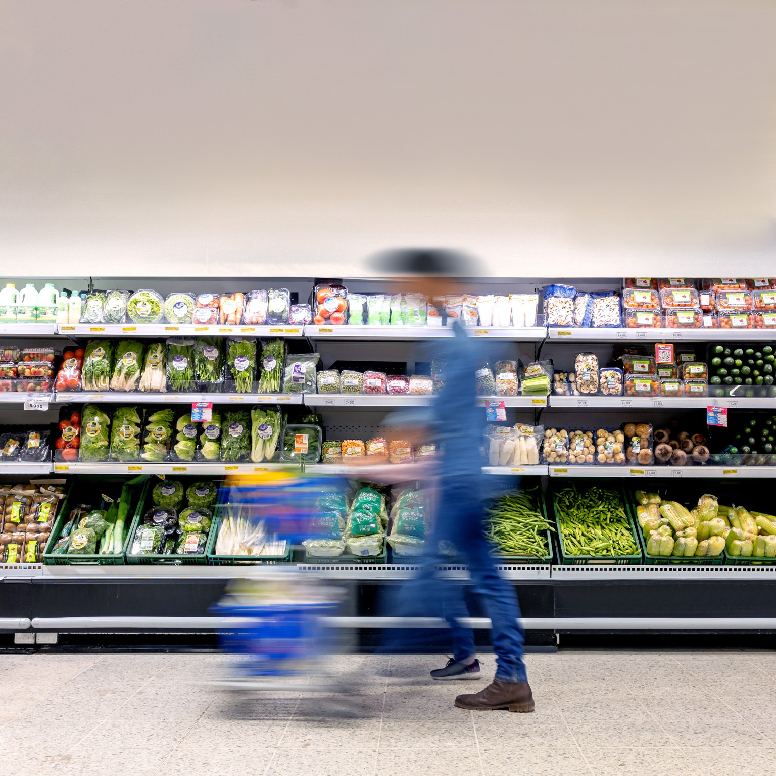 A man maneuvering a shopping cart through the grocery store, passing by the refrigerated produce section.