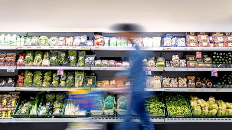 A man maneuvering a shopping cart through the grocery store, passing by the refrigerated produce section.