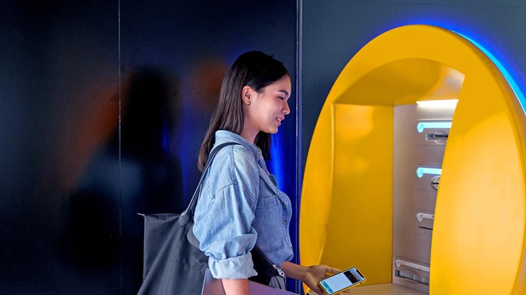 A woman with a smile on her face is standing at an ATM, holding a cell phone and laptop, as she prepares to withdraw money.