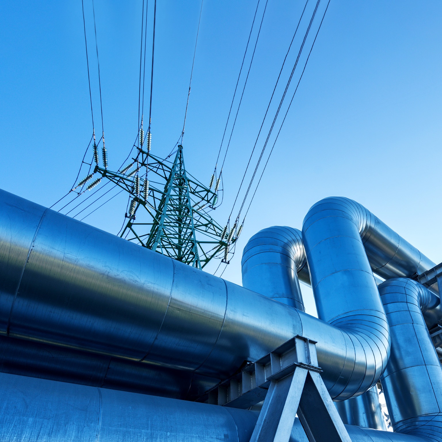 Large metal pipes tansporting oil curve and ascend against a clear blue sky with a tall electricity pylon visible in the background.