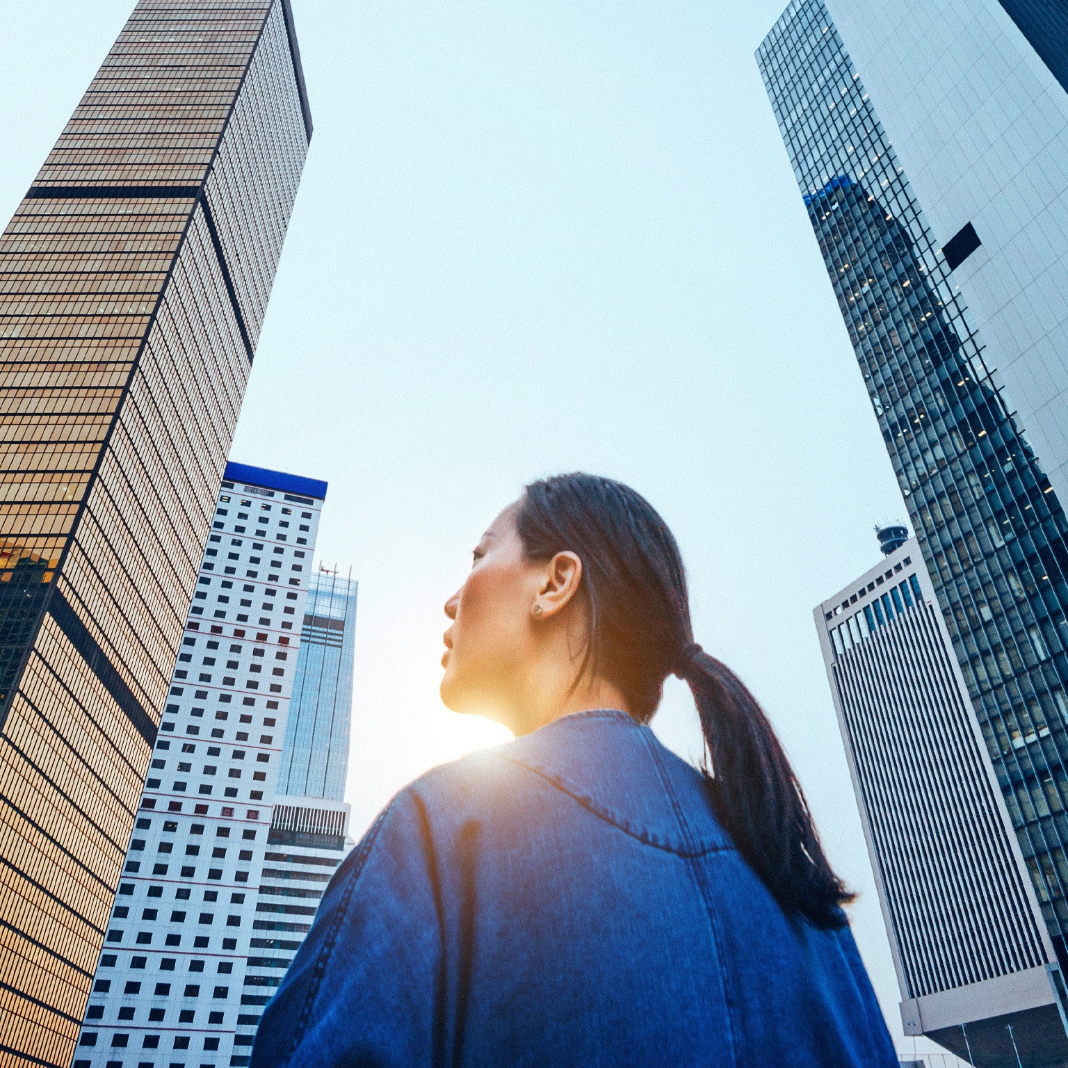 A woman stands with her back to the camera, gazing upward at a cluster of towering skyscrapers that dominate the frame. 