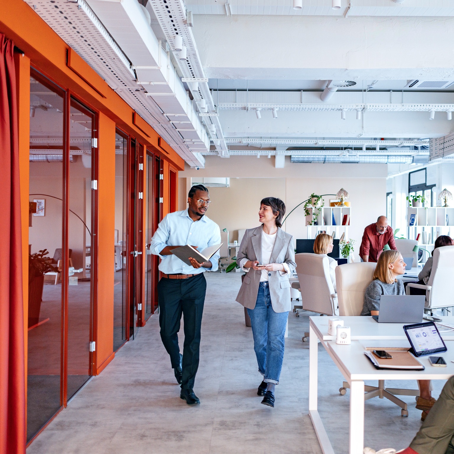 A bustling, modern office space with a diverse group of employees engaged in various work activities, some collaborating in groups, while others work individually at their desks. Two colleagues are seen walking and talking in a hallway, showcasing a dynamic and collaborative work environment. 