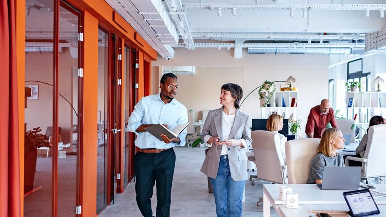A bustling, modern office space with a diverse group of employees engaged in various work activities, some collaborating in groups, while others work individually at their desks. Two colleagues are seen walking and talking in a hallway, showcasing a dynamic and collaborative work environment.