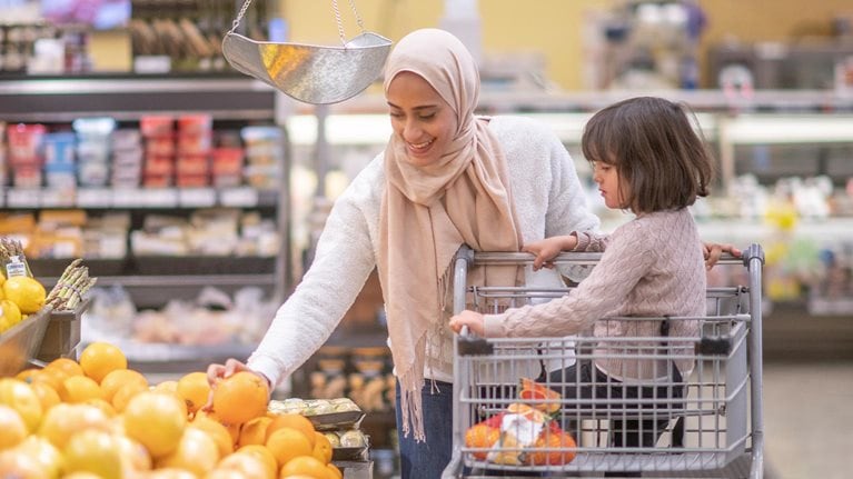 A Muslim mother goes grocery shopping with her daughter. Her daughter is sitting on the grocery cart while she picks through some fresh produce.