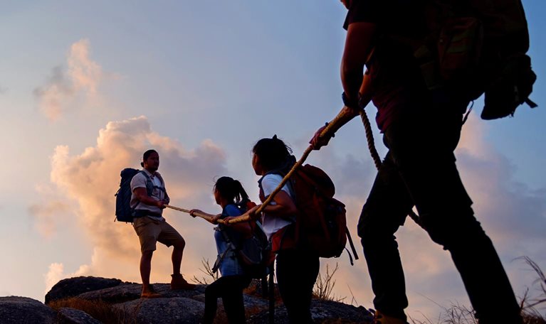 Group of friends hikers climbing up silhouette mountain cliff.