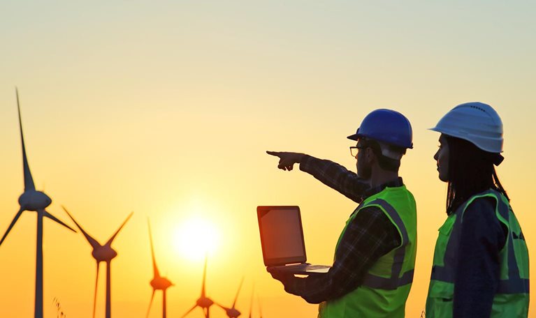 Two workers in a Windmills field working on a laptop and pointing in the direction of the windmills as the sun is setting. 