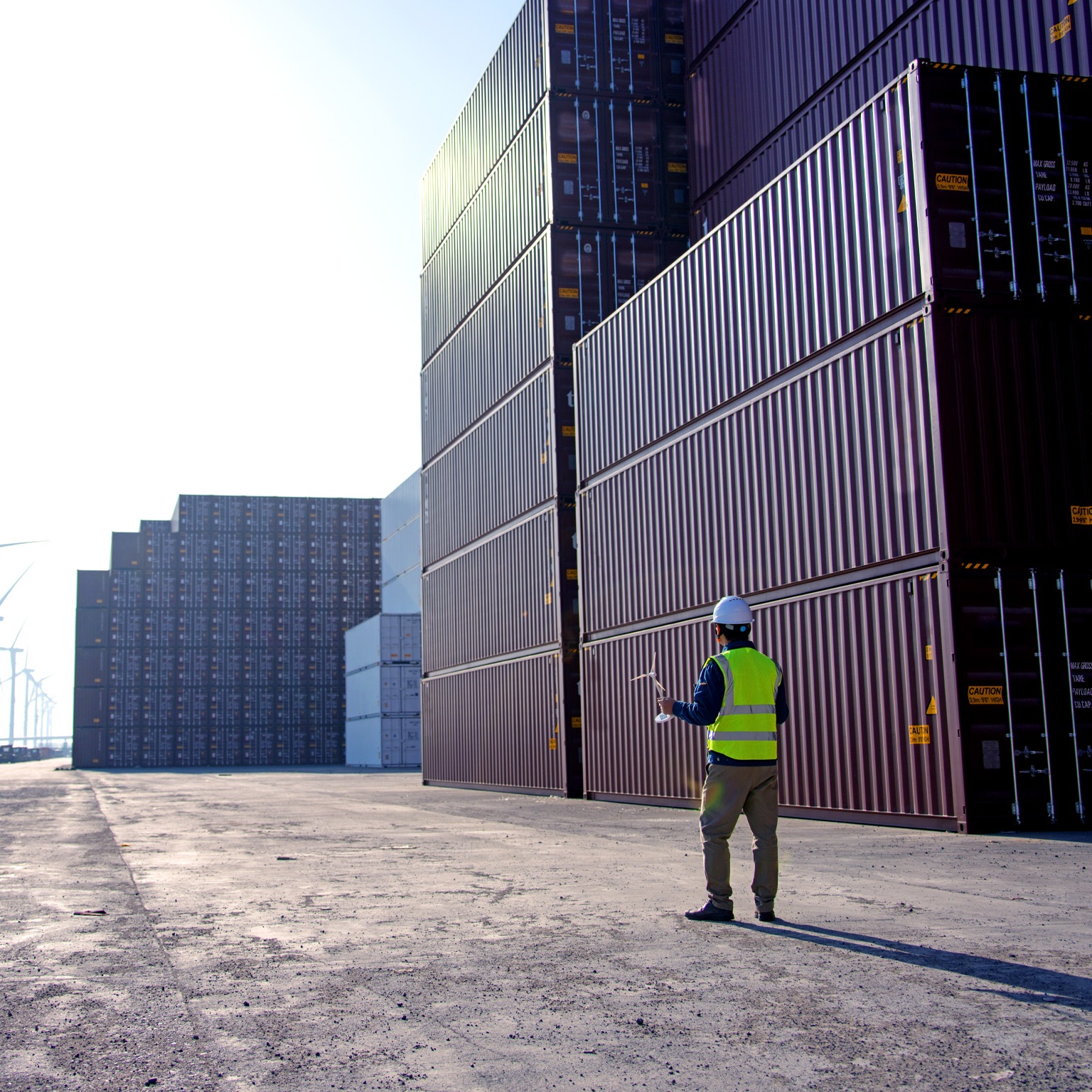 A worker in a dockyard, donning a helmet and reflective vest, gazes at a blueprint of a windmill. In the distance, there are rows of trucks and wind turbines.