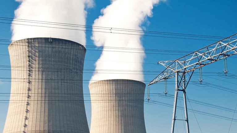 Two large cooling towers at a power plant releasing thick white steam into a clear blue sky, with high-voltage transmission lines and electrical towers in the foreground.