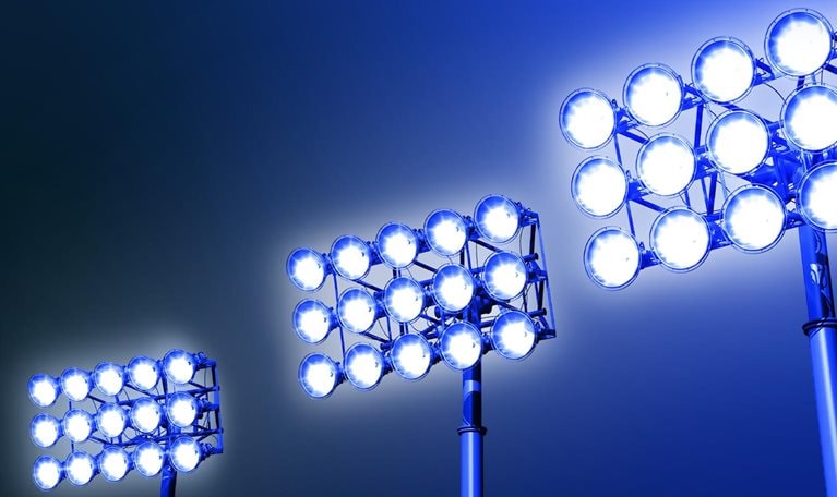 Against a dark blue backdrop, three sets of stadium lights emit a radiant glow, evoking a feeling of excitement and suspense.