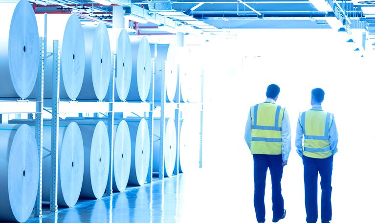 Workers in reflective clothing walking along large paper spools in printing plant