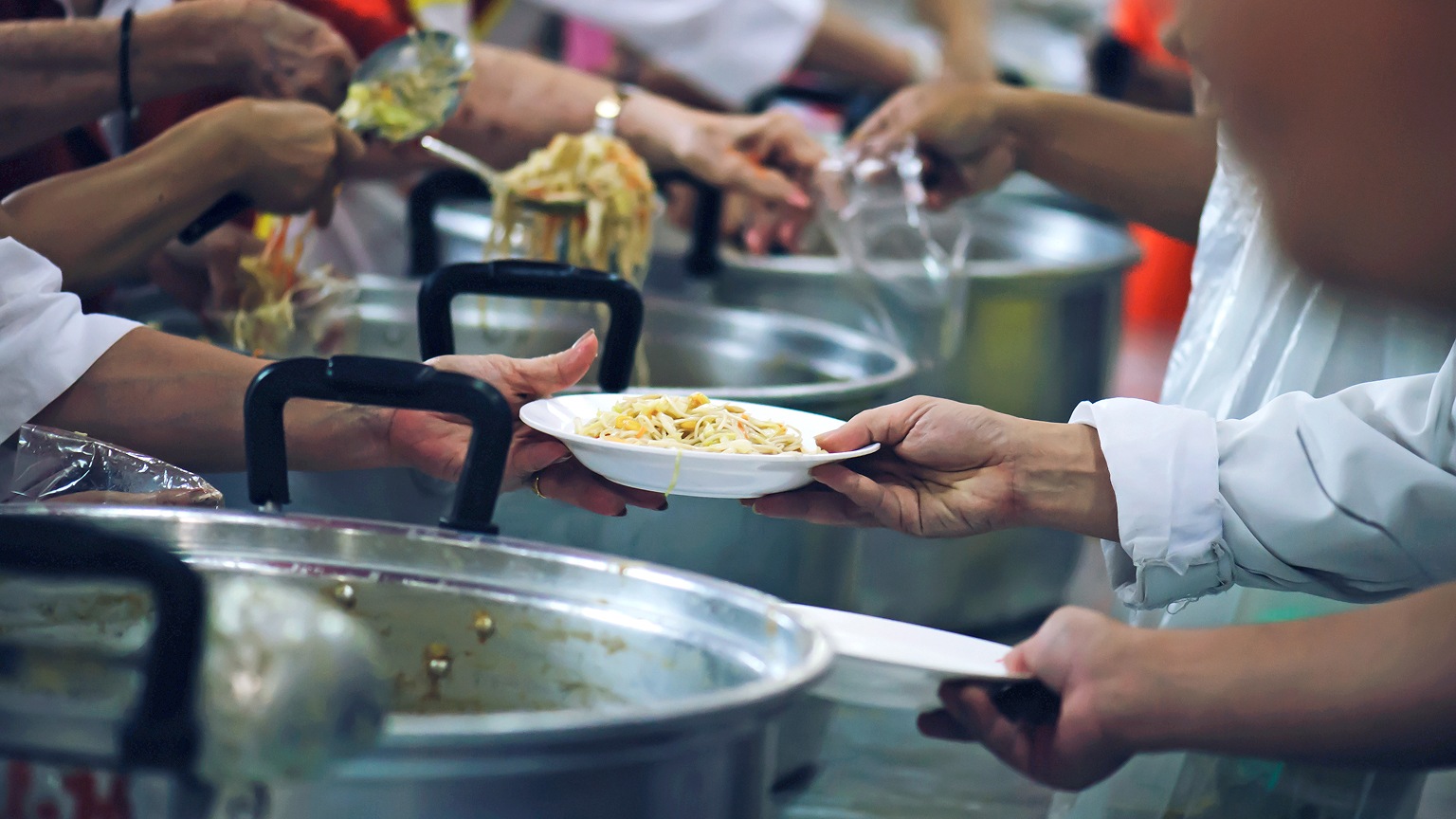 A close-up of several people serving food in a communal setting, with hands passing a white plate of noodles over large metal pots while others scoop servings using ladles.