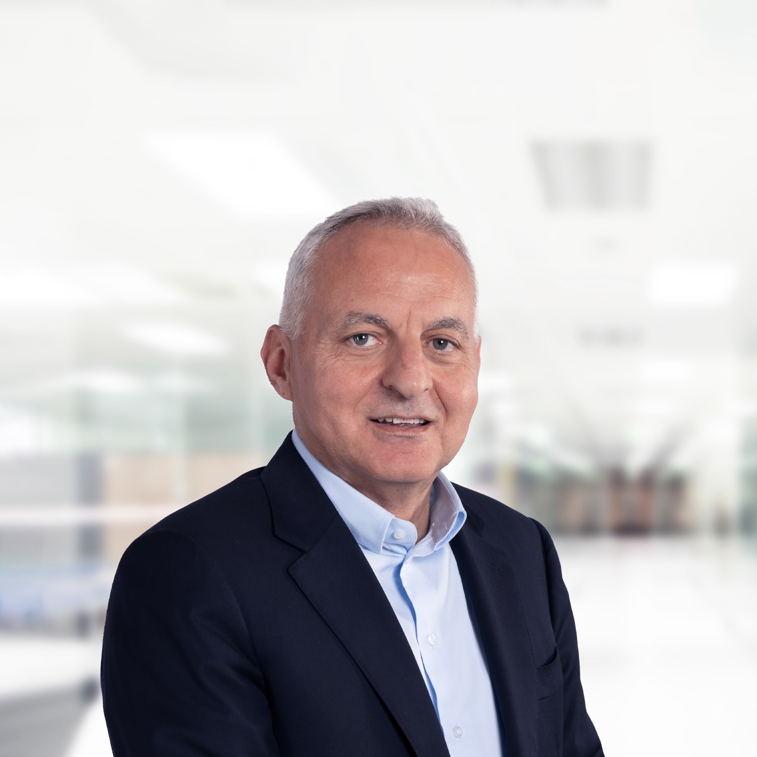 Tufan Erginbilgiç is depicted in a navy blue suit jacket and light blue button-down, smiling against a bright, blurred office setting.