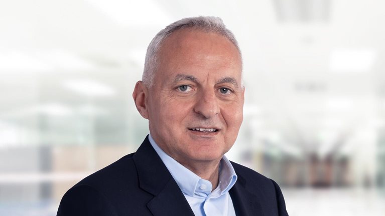 Tufan Erginbilgiç is depicted in a navy blue suit jacket and light blue button-down, smiling against a bright, blurred office setting.