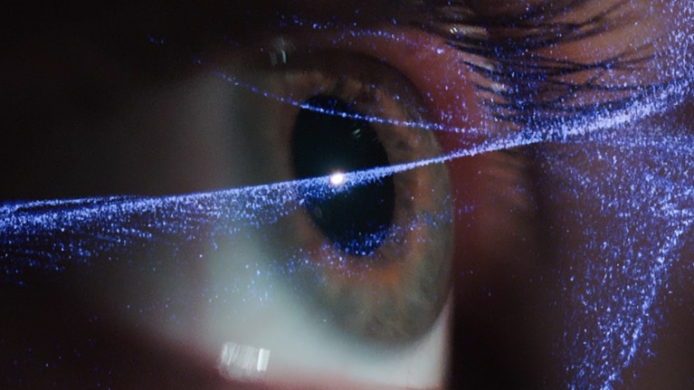 Close-up of a human eye, with glowing blue, digital particles swirling across the pupil and iris.