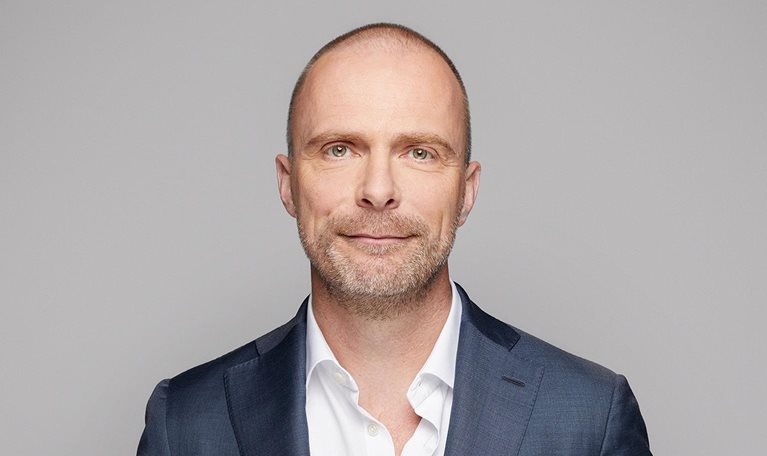A portrait of Robert Carsouw, with a light beard, and a easy smile. He is wearing a dark blue suit and a white, open-collared shirt against a light gray background.