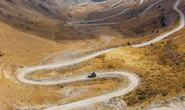 A silver SUV makes navigates a winding dirt road as it snakes through a wide mountain valley, cutting sharp switchbacks across dry, golden-brown slopes. Steep ridges rise on both sides, with sparse vegetation and distant peaks fading into a hazy, expansive backdrop.
