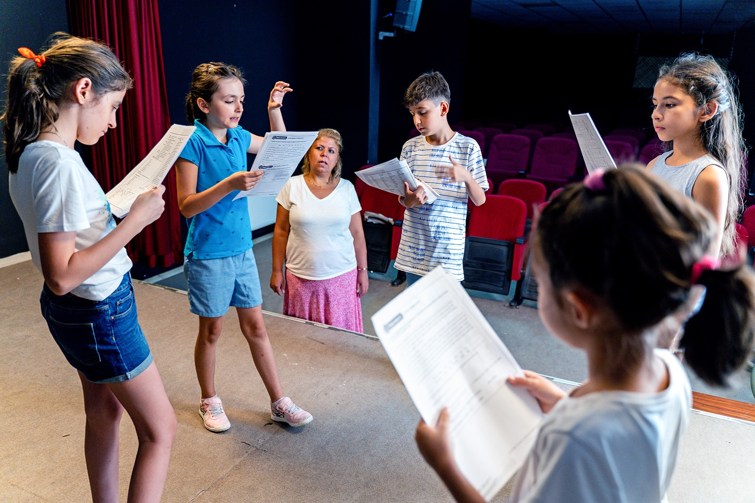 Group of children reading scripts on a stage while their drama teacher looks on.