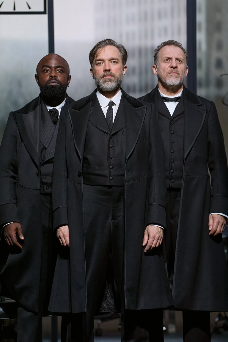 Three members of the The Lehman Trilogy standing defiantly and dressed in black vintage tuxedos and overcoats at the National Theatre. Photo by Mark Douet.