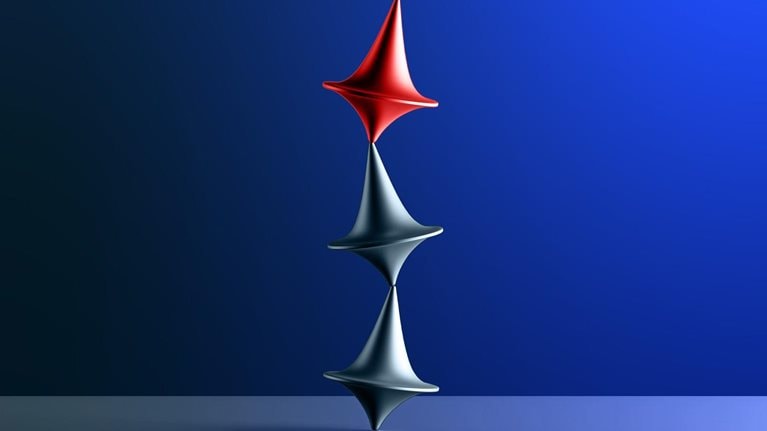 Three tops, two silver and one red, are arranged in a vertical stack on a blue surface. The tops are uneasily balanced, with the red top positioned above the two silver ones.