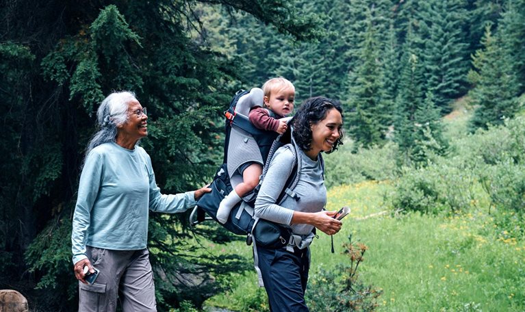 A smiling family, composed of a grandmother, grandfather, mother with a baby in a carrier on her back, and a young girl, treks through a forest, crossing a wooden bridge over a small stream. The grandfather assists the girl as they walk.