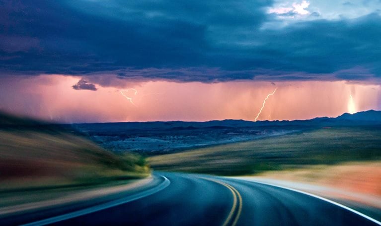 A dark asphalt road curves through a motion blurred landscape beneath a dramatic, stormy sky. In the distance, lightning flashes behind rain clouds, illuminating the horizon with an eerie light.