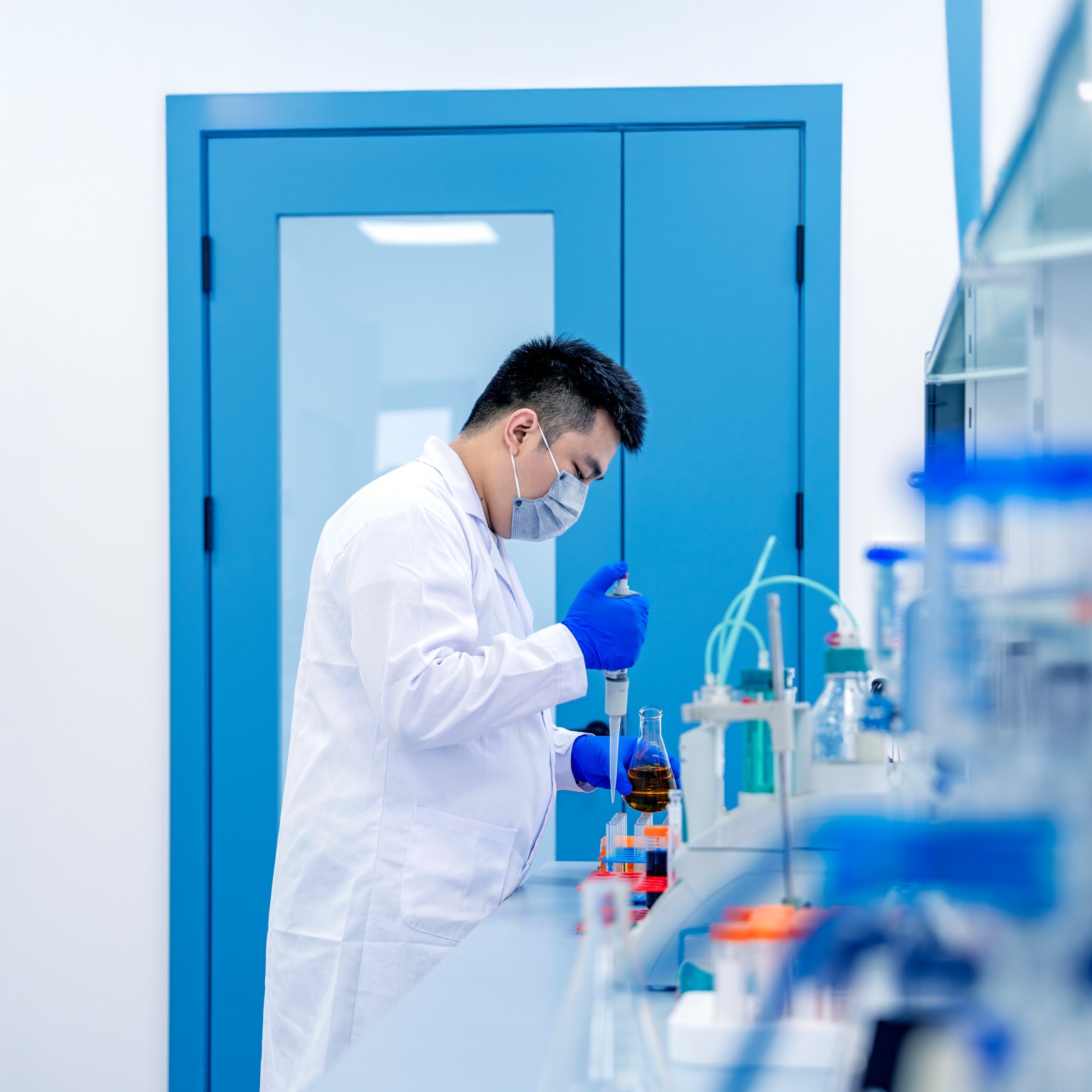 A lab researcher wearing a face mask, gloves, and a white coat is using a pipette to transfer liquid into a flask on a laboratory bench. The scene shows a clean, modern lab environment with scientific equipment and containers arranged for experimental work.