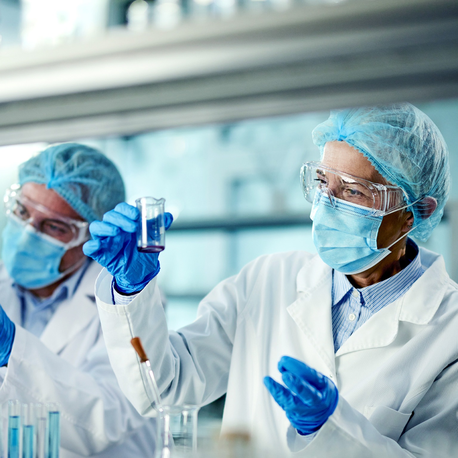 Two scientists in a laboratory are wearing protective gear including lab coats gloves and face masks One scientist carefully examines a liquid in a beaker while the other holds test tubes containing samples.
