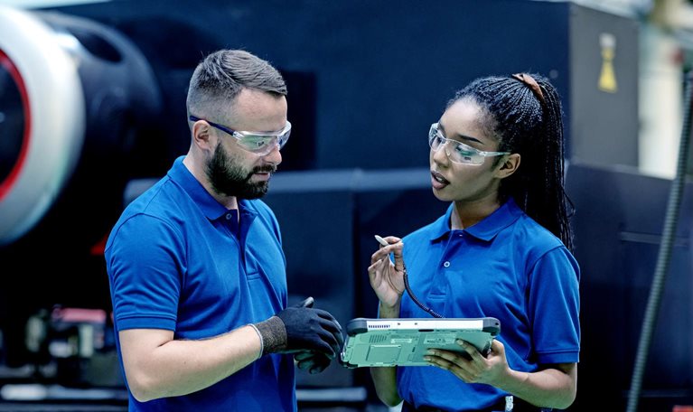 In an industrial manufacturing setting, a supervisor and process engineer, both dressed in blue company shirts and wearing protective glasses, engage in a conversation while examining a tablet near a sizable machine.