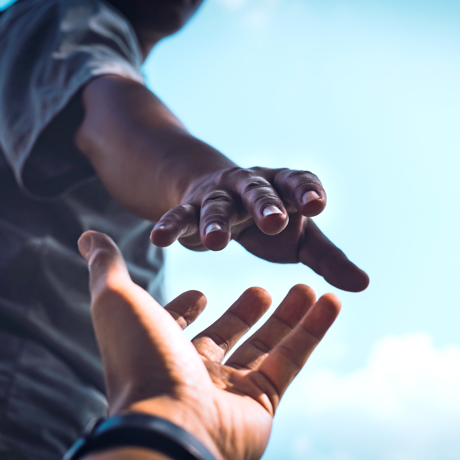 A tightly cropped image of a person reaching down with an outstretched hand while another person reaches upward, their fingers almost touching. Set against a bright sky, it conveys a strong sense of help, connection, and support.