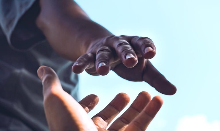 A tightly cropped image of a person reaching down with an outstretched hand while another person reaches upward, their fingers almost touching. Set against a bright sky, it conveys a strong sense of help, connection, and support.