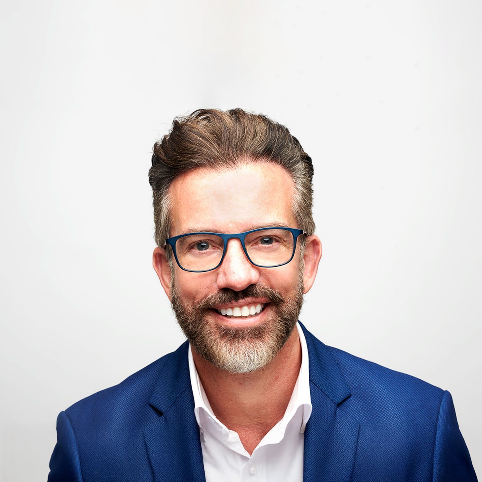 Nick Jeffery smiling warmly at the camera, wearing glasses and a blue suit jacket over a white shirt. He has neatly styled brown hair and a trimmed salt-and-pepper beard, set against a clean, light background.