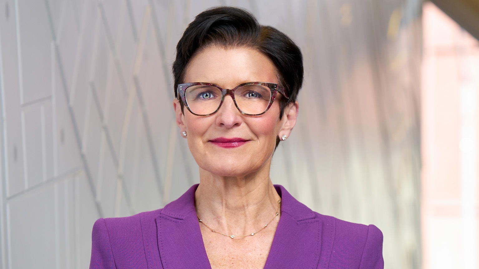 A portrait of Jane Fraser with short dark hair wearing glasses and a purple blazer over a black top. She stands against a softly blurred, modern interior background and looks directly at the camera with a confident, composed expression.