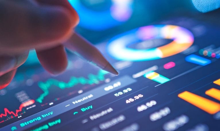 A hand uses a stylus to interact with a tablet displaying financial data and charts, including candlestick charts and pie charts. The screen glows with blue and orange hues.