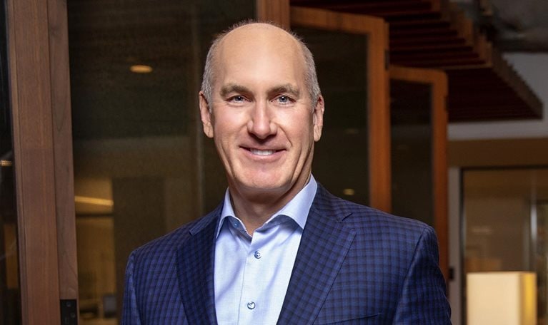 A portrait of John Stankey smiling, wearing a royal blue suit jacket over a blue button-down dress shirt.