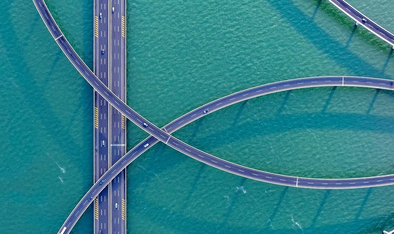 An aerial view of sweeping, elevated highways curving gracefully over turquoise water, creating an intricate, looping pattern. A few cars travel along the roads, adding scale and movement to the striking geometric scene.