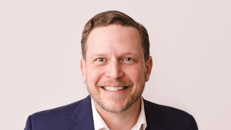 Nate Boaz, wearing a white button-down shirt and blue jacket, is captured with a smile on his face against a pale tan background